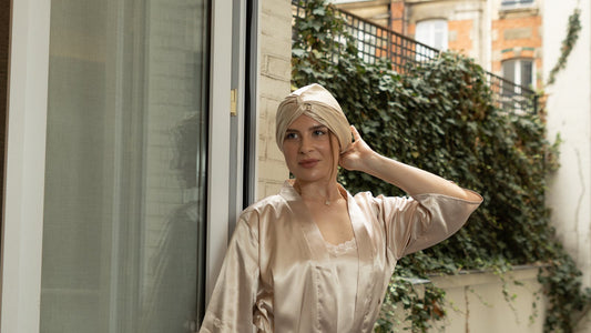 A woman at an open French window in Paris, adjusting a Gold Siluxe Glow silk bonnet with its twist knot at the front