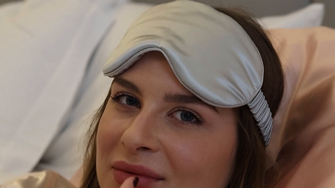 A woman lying on a Rose gold silk pillowcase looks at the camera, a Silver silk eye mask pushed up onto her forehead