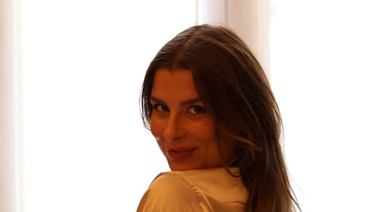 A woman at a window with sheer voile curtains looks back over her shoulder and smiles, her long brown hair worn loose