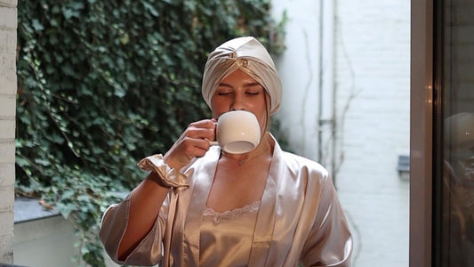 A woman sips coffee with her eyes closed, wearing a Gold Glow silk bonnet with its twist at the front and a Gold silk scrunchie on her wrist