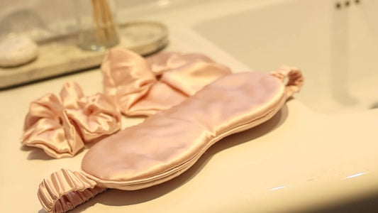 A Rose gold silk eye mask and two Rose gold silk scrunchies laid on a white bathroom counter beside the basin