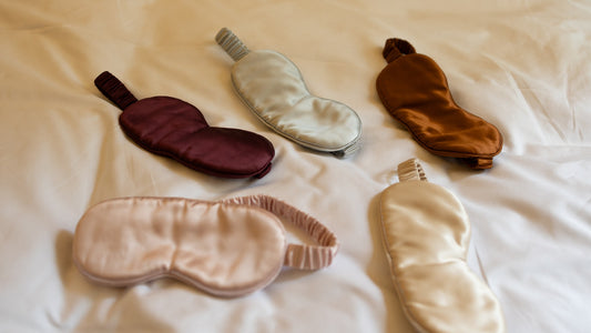 Five silk eye masks in Burgundy, Silver, Bronze, Rose gold and Gold laid out on a white duvet