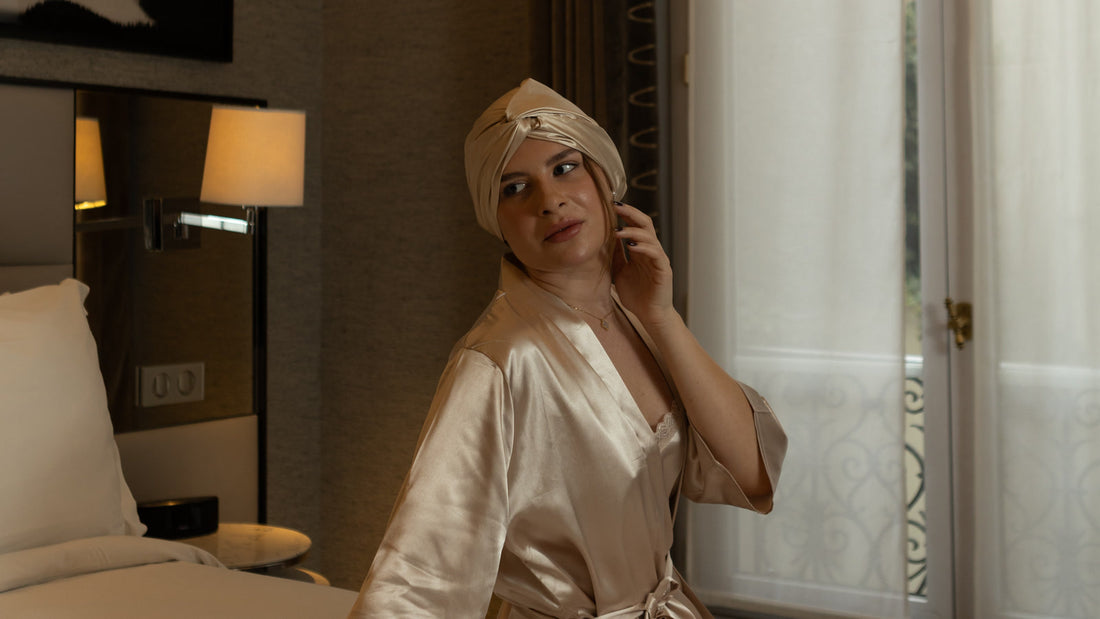 A woman in the Gold Glow silk bonnet and a champagne satin robe glances over her shoulder in a hotel room, a lamp behind her and a tall sheer-curtained window