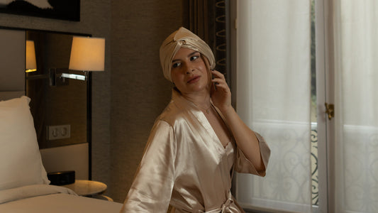 A woman in the Gold Glow silk bonnet and a champagne satin robe glances over her shoulder in a hotel room, a lamp behind her and a tall sheer-curtained window