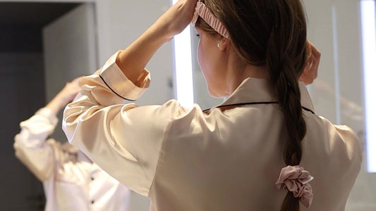 Seen from behind at a bathroom mirror, a woman in satin pyjamas slides on a Rose gold silk headband, her loose braid tied with a Rose gold silk scrunchie