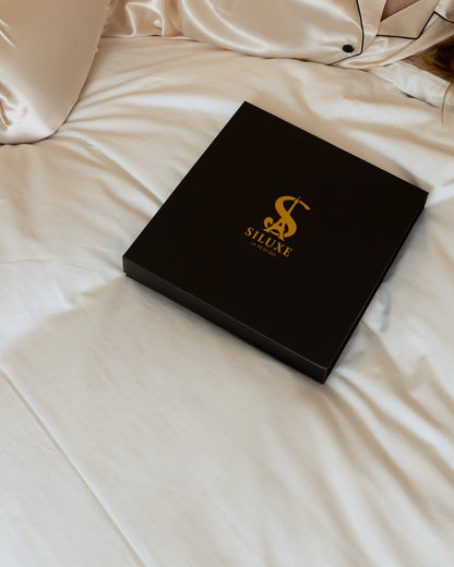 The black Siluxe gift box, closed, gold S monogram, resting on the bed